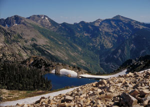 A rocky hillside makes the foreground, marking out a lake in the forested valley beyond.  Far off, a series of mountains rises up to touch the sky.