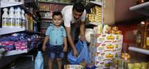 In a store aisle, a young boy looks into the camera while his father packs food into plastic bags.