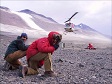 Two men crouch as a helicopter lands in the McMurdo Dry Valleys of southern Victoria Land.