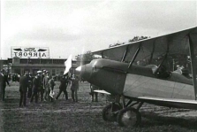 One of the two Swallow aircraft in the 1925 National Air tour, equipped with a Curtiss OX5 engine, is shown departing Ford Airport. Edsel B. Ford, one of two official starters for the 1925 tour, can be seen with the large starting flag.
