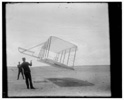  Side view of glider flying as a kite near the ground, Wilbur at left and Orville at right, glider turned forward to right and tipped downward.
