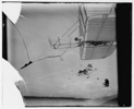  Side view of Wilbur Wright piloting a glider in level flight almost overhead, moving to left, showing bottom wing and elevator.
