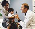 A mother holding her baby in a doctor's office; the doctor is sticking his tongue out to make the baby laugh.
