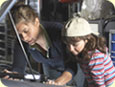 A female mechanic and a little girl looking under the hood of a car.