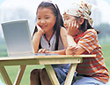 Two little girls, sitting outside,  looking into a laptop.
