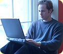 A man sitting by a large window, looking at his laptop.