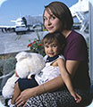 A mom, son and his teddy bear sitting by a big window at the airport; an airplane is in the background.