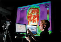 SNL researcher Rob Abbott uses a joystick and plays the role of a student in a training exercise driving an amphibious assault vehicle simulator used by the Navy and Marines. The second monitor is an instructor/operator application called CDMTS. In the background is a thermal image of a student's face used for investigating biometrics to monitor the student in various ways, including the level of engagement and focus of attention. Photo by Randy Montoya.