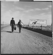 Image shows two persons walking (hitchhiking) along road near billboard