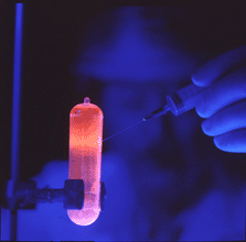 Under blue light, researcher uses a syringe to extract purified DNA, which glows orange, from a tube.