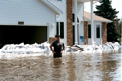 Man standing in front of new home in water 
up to his hips. House protected by sandbags.
