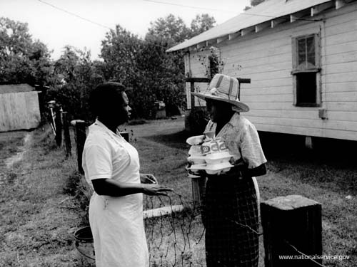 When VISTA Alice Fay White, 23, of Opelousas, Louisiana, originally arrived in Little Rock in 1972, there was no assignment for her, so she began to seek funds and support for a senior citizen hot lunch program in Sweet Home, Arkansas. She got her funding through a Community Action Program emergency food and medical program grant, and she became the "coordinator" to keep the program going, working in the kitchen preparing food and delivering the lunches. In this photo, a staff member delivers lunch to an elderly woman in Sweet Home.