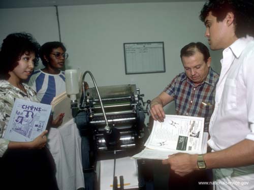 Printer Isidro Garcia (plaid shirt) discusses the publication of the next issue of CHINS Notes, a magazine reflecting the experiences of young people in Las Cruces, New Mexico, with VISTA members (left to right) Annette Alvarez, Norma Scott, and Derek Rubio in 1986.
