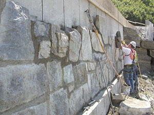 Masonry workers are attaching a historically compatible stone veneer to a new concrete retaining wall near Triple Aches.