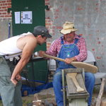 A craftsman explains to a visitor how he makes boat paddles by hand.