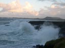 Waves crash on a rocky beach around sunset.  White water sprays off the black rocks, with pastel sunset colors lighting the sky in the background