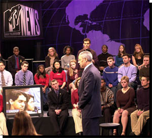Secretary Powell standing before an audience of young people, looking at TV monitor showing young man in India asking question of the Secretary