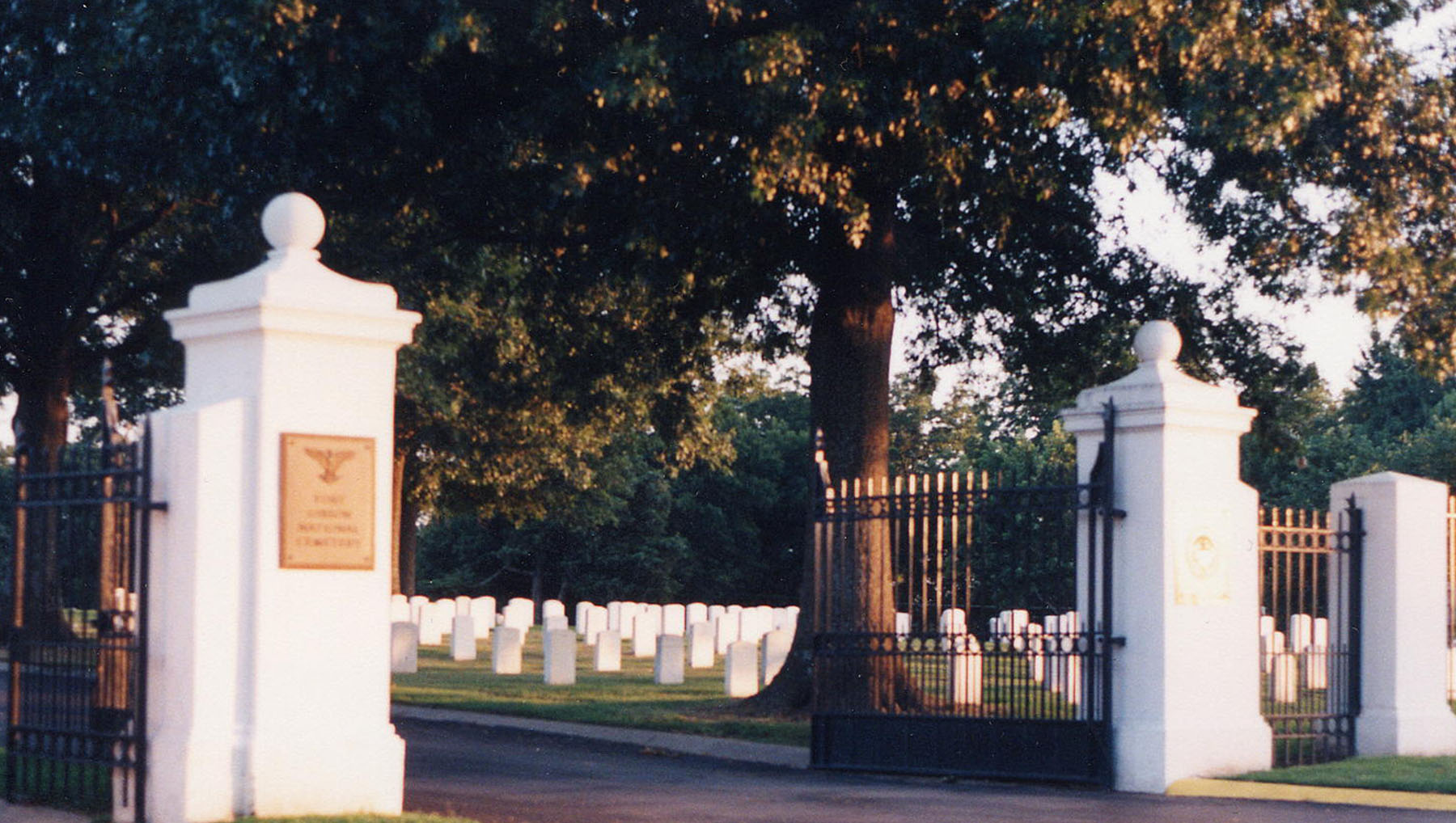 Photo of an opened iron gates attached to pillars leading into the cemetery with upright headstones in the background.