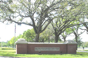 A photo of a brick wall structure with the name Biloxi National Cemetery engraved on front.  Two large trees, the American Flag, and upright markers are shown in the background.