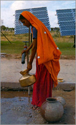 Photo of a woman filling a gourd with water at a pump that is in front of a PV module. 