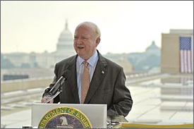 Photo of a balding man in a suit standing at a podium with the DOE seal on it. Stretched out on the roof behind him are a number of solar power panels, and a U.S. flag hangs behind that. The U.S. Capitol Building is in the distant background.