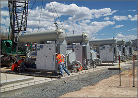 Photo of several men wearing hardhats and safety vests and guiding a modular power unit into place as a crane holds it inches above its foundation. Four other power units are already in place in a line beside it, each consisting of a cylinder about 2 feet in diameter and 15 feet long, mounted atop an electrical equipment box about six feet square and four feet deep. The end of another large cylinder sticks out behind the electrical box and has a 6-inch pipe running from it.