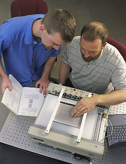 Photo of NIST researchers John Roberts and Oliver Slattery using the tactile graphic display device to depict the NIST logo.