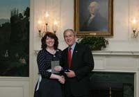 President Bush presents the President's Volunteer Service Award to Army spouse Colleen Saffron from Fort Hood, Texas, before the White House celebration of  Military Spouse Day on Tuesday, May 6, 2008. Saffron's husband, Staff Sergeant Terry Saffron, was severely injured in Iraq in May 2004 when an improvised explosive device was detonated, shattering his lower right jaw and injuring his right arm, resulting in loss of all muscle tissue.  In March 2007, Saffron collaborated with a Navy and Air Force spouse to create Operation Life Transformed.  This non-profit charitable foundation helps educate and train military spouses and caregivers of wounded troops, which allows them to work from home while caring for their loved one.  To date, Operation Life Transformed has paid for 30 spouses and caregivers to enter online training programs.  Saffron, who is legally deaf, also volunteers with Texas Hearing and Service Dogs, an organization that trains dogs, free of charge, to assist people living with deafness or mobility-related issues.  Saffron and her hearing dog, Gretchen, visit schools to educate children about how service dogs can assist the disabled.