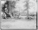  Katharine Wright lying on the ground, covered by a blanket, her friend Harriet Silliman in a chair nearby, in wooded camp area. 
