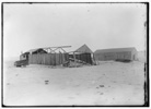  View of the camp at Kitty Hawk from the northeast, showing its condition at the time of Wilbur's arrival on April 10, 1908. The old 1902 building is on the left, its side walls still standing but its roof and north end gone. The remains of the 1902 glider are on the ground.
