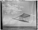  Left side view of the 1900 Wright glider before installation of forward horizontal control surface, flying as a kite, tipped forward; Kitty Hawk Lifesaving Station and Weather Bureau buildings in background to the left. 


