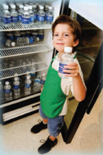 Boy holding a bottle of water standing in front of an open refrigerator filled with bottled water.