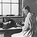 A 1937 NIH laboratory technician surrounded by tools of the trade; a rack of cotton-stoppered test tubes, a microscope and various glass jars.