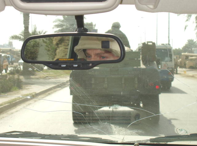 Photograph taken from inside a vehicle looking out through the cracked windshield at the military vehicle in front.  In the rearview mirror you can see the driver’s eyes and the front part of his/her helmet. 