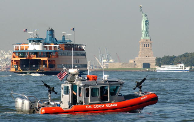 A photograph of a U.S. Coast Guard boat in the foreground with a ferry  and the Statue of Liberty in the background.