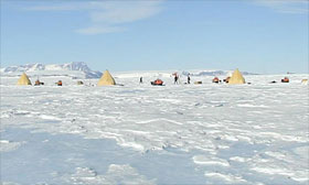 Each pyramid-shaped Scott tent housed two people. Snowmobiles, food boxes, solar panels, wind turbine, radio antennas, people, snow, and blue ice complete the picture.