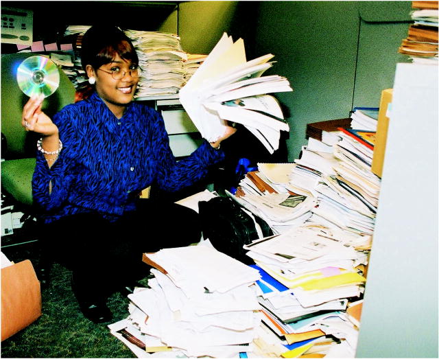 Photo shows a Department of Education employee, surrounded by stacks of existing Federal K-12 education paperwork, holding a computer disk.