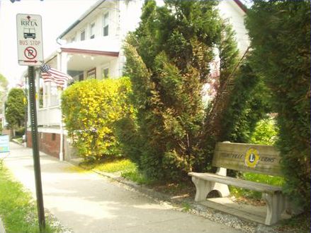 The second picture shows a bus stop with a sidewalk and a bench between two trees. The bench was placed there by the Lion’s Club. There is a bus stop sign in the grass strip between the sidewalk and the curb.