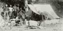 Univ. of Washington historic photo: Several men stand near a canvas tent with various camp objects on the ground around them