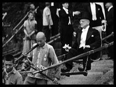 A man in morning suit is carried in a sedan chair by three Chinese men, surrounded by other men in formal dress