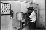 Man drinking at a water cooler in the street car terminal; sign above it reads: 'Reserved for Colored'