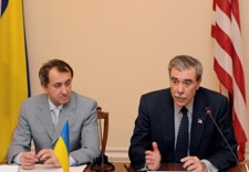 Gutierrez and Danylyshyn sit at a table in front of microphones and are flanked by United States and Ukraine flags.