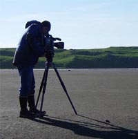 A man shooting video footage on a beach on St. Paul Island with grassy tundra in the background.