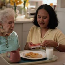 Fotografía de una mujer mayor almorzando con la asistencia de su cuidadora