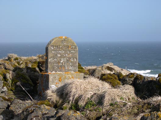 A weathered, lichen-covered concrete headstone sits atop a hill at Northeast Point, with the Bering Sea in the background