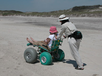Woman pushing another person sitting in large-tired beach wheel chair.