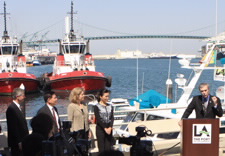 Gutierrez on podium surrounded by press with Port harbor and ships in background. Click for larger image.