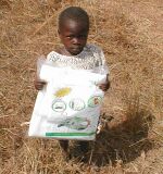 Little girl standing in a field, holding a plastic bag (that is nearly as big as her) that contains a new insecticide-treated bed net