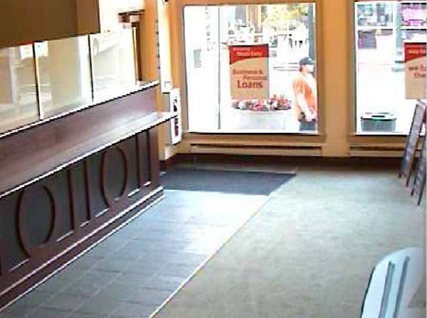 A surveilance camera photo of the front / right side of the male suspect. Suspect is wearing an orange tshirt, dark pants, and a baseball cap. Photo was shot through a storefront window.