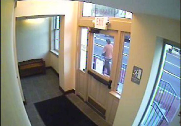 A surveilance camera photo of the rear of the male suspect. Suspect is wearing an orange tshirt, dark pants, and a baseball cap. Photo was shot through a doorway window.